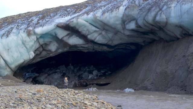 A person standing outside an ice cave near a riverbank with rocky terrain.