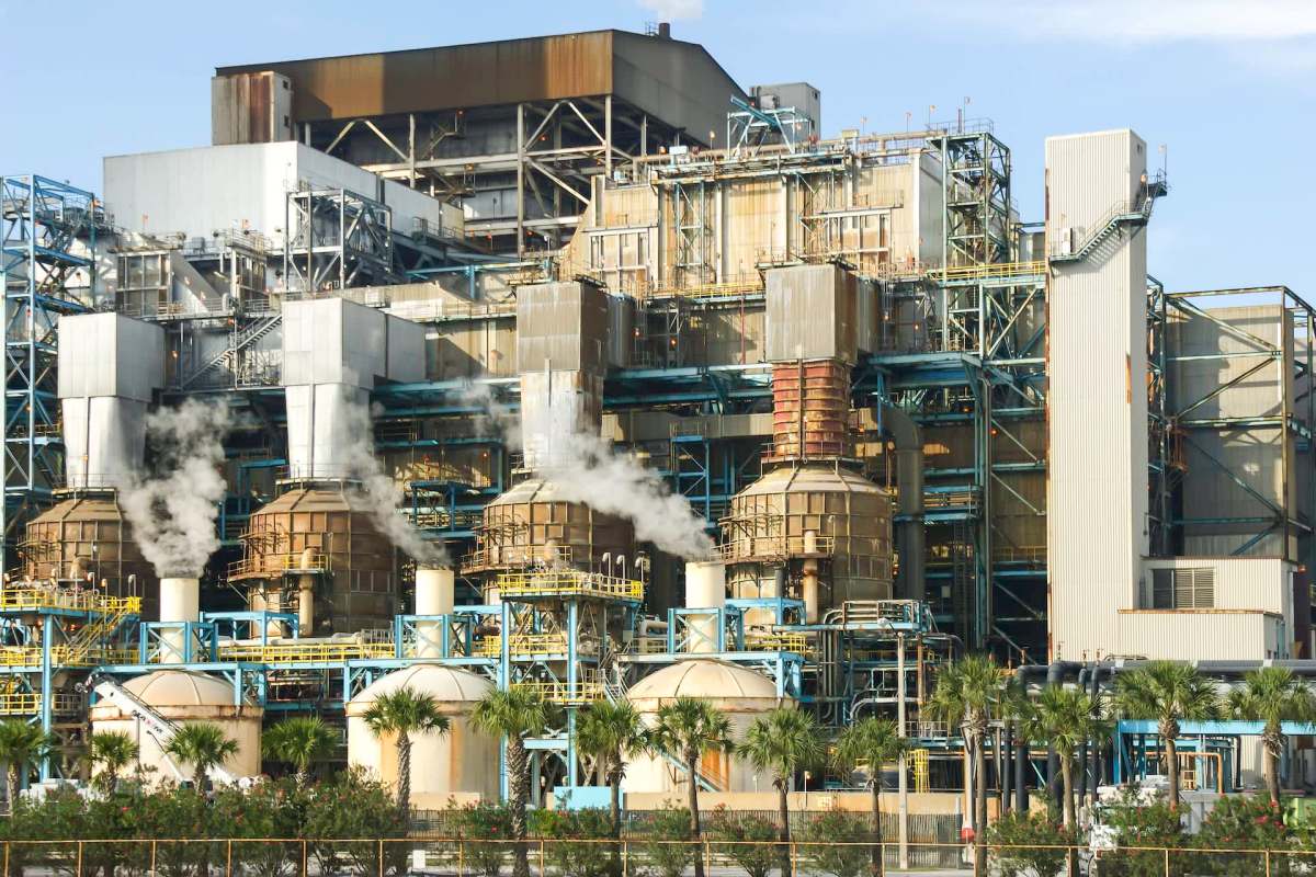 A large industrial facility with smoke rising from multiple structures and palm trees in the foreground.
