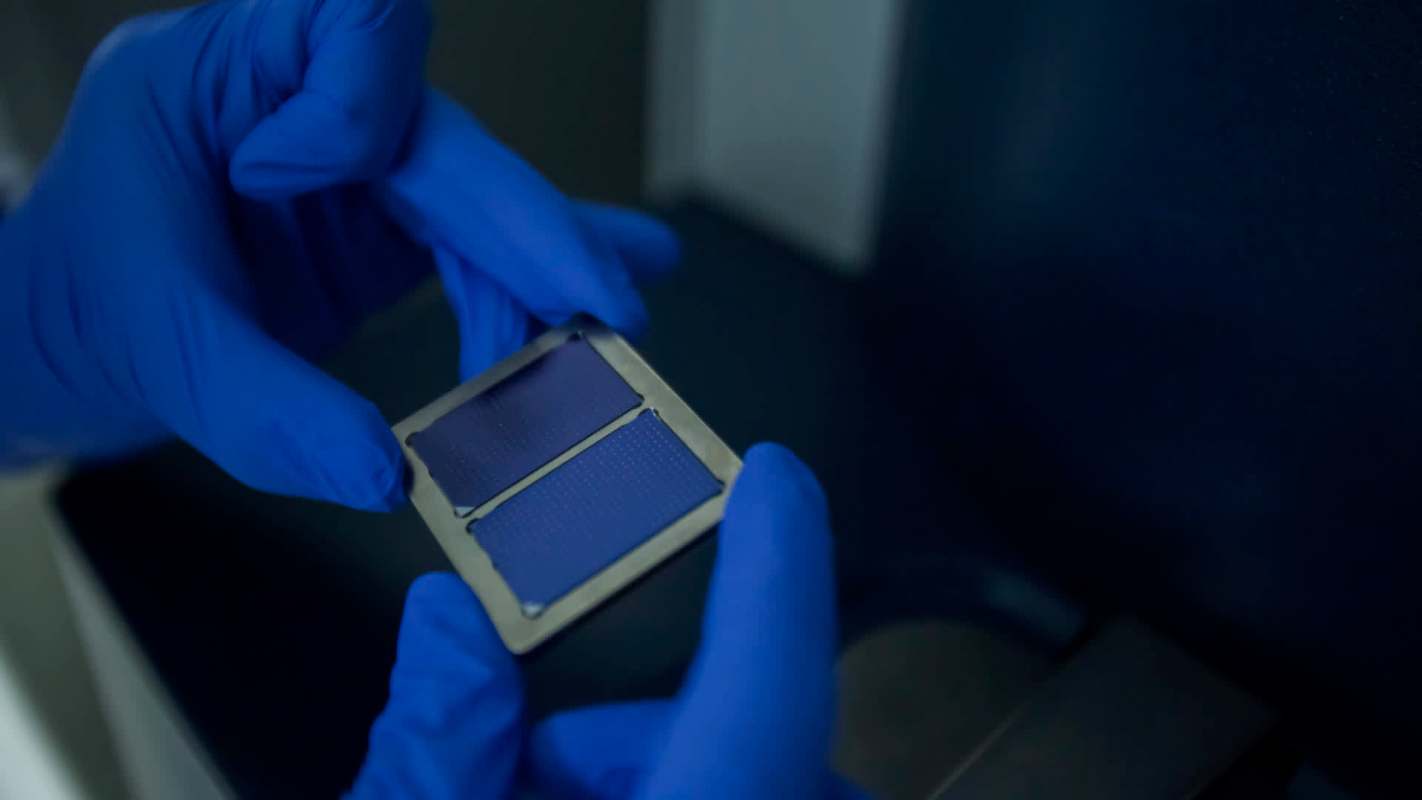 A person in blue gloves holds a small rectangular device with a dark surface, possibly a solar cell or semiconductor.