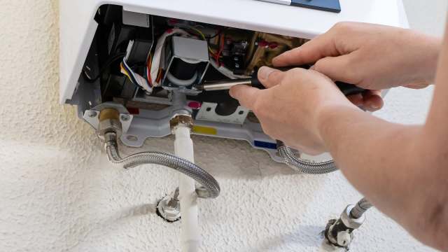 A person repairs a water heater, using a screwdriver in a tight space with visible pipes and wires.