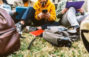 Students sitting on grass with school supplies and a phone.