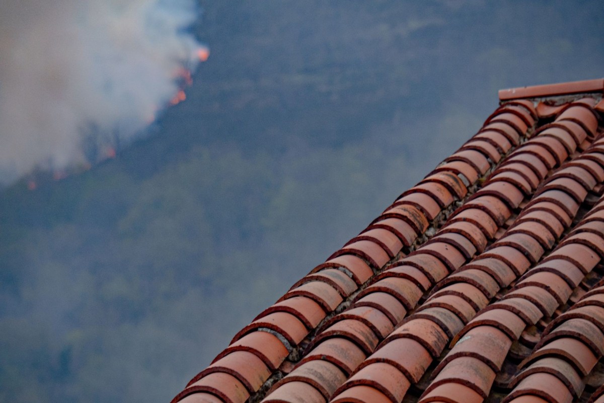 The side of a terracotta roof with a wildfire in the mountain off in the distance
