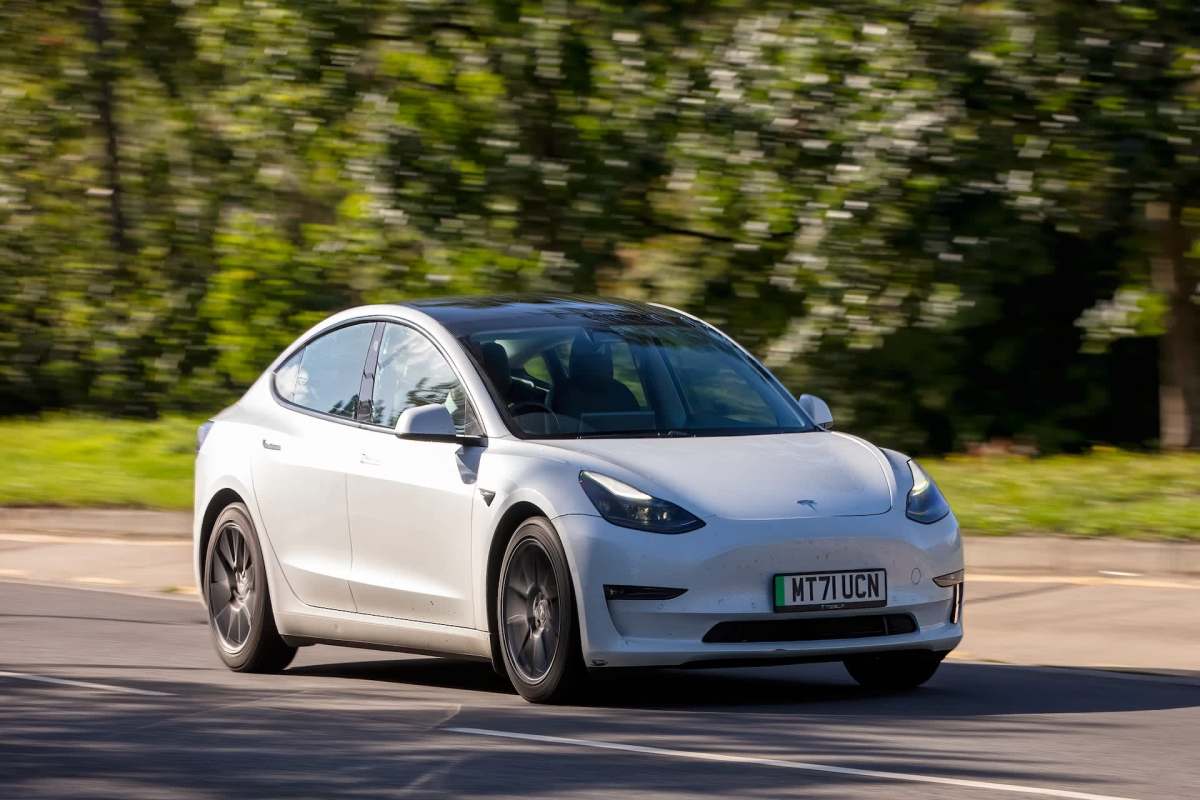 A white Tesla Model 3 drives along a road with trees in the background.