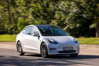 A white Tesla Model 3 drives along a road with trees in the background.