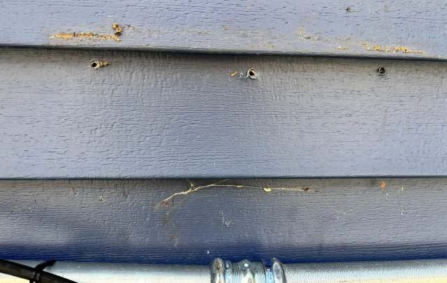 A blue house siding with two small holes and some dirt or debris near a metal pipe.