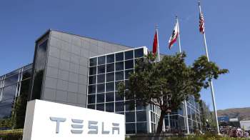 Tesla building with a large sign, flags flying, and a tree in front.