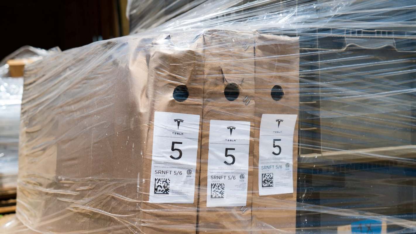 Boxes labeled with the Tesla logo and number five, wrapped in plastic on a pallet.