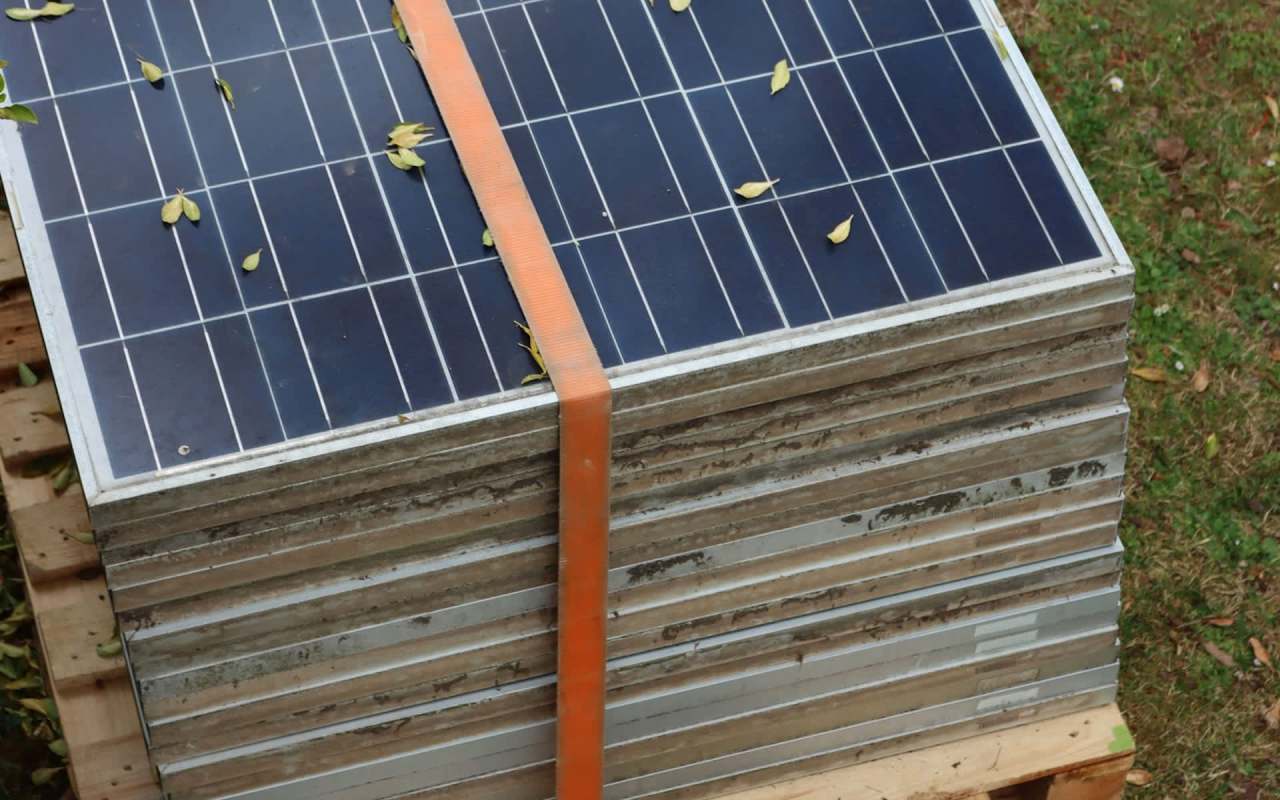A stack of solar panels secured with an orange strap, placed on wooden pallets with scattered leaves around.