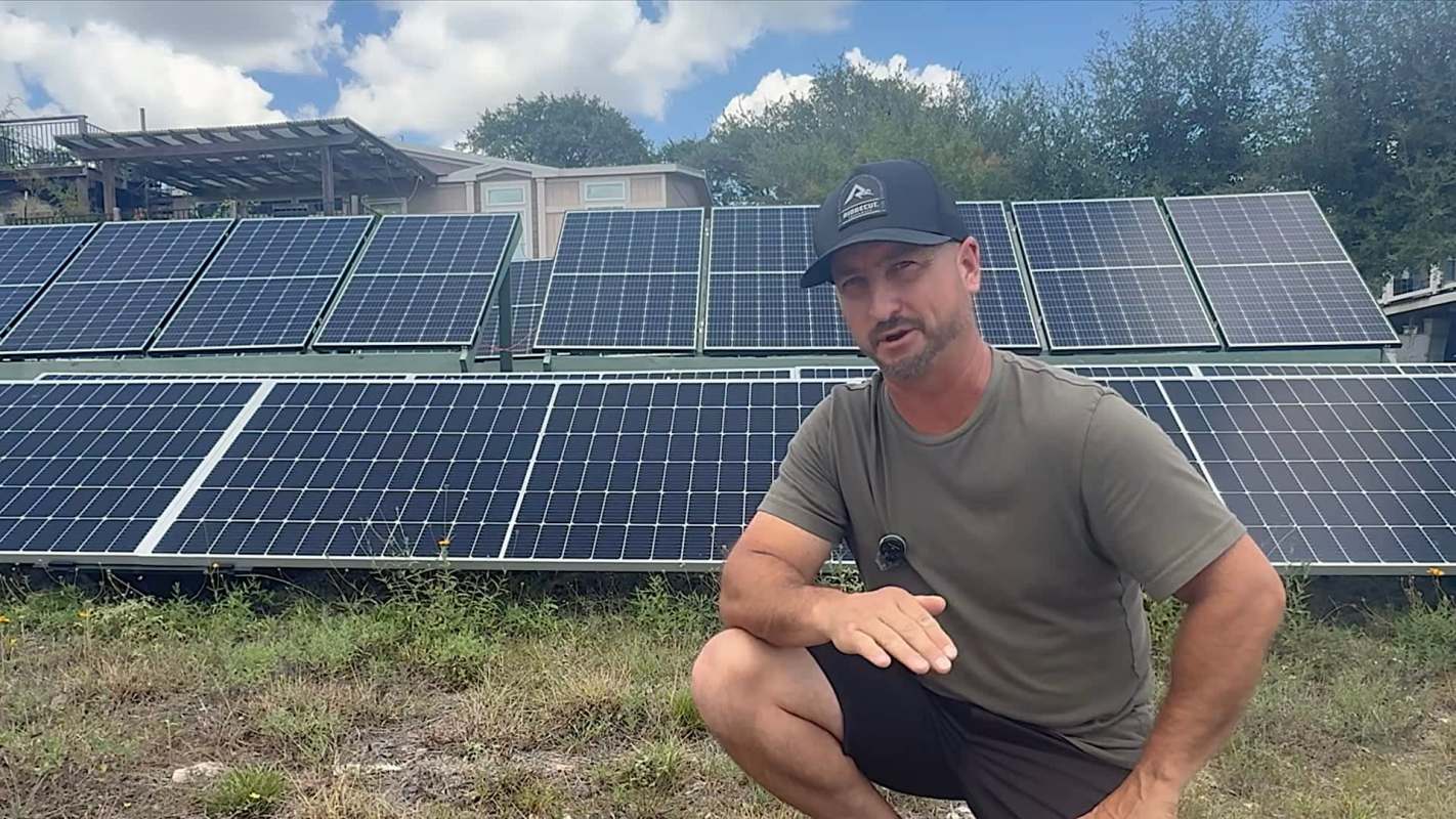 A man kneels in front of solar panels in a grassy area.