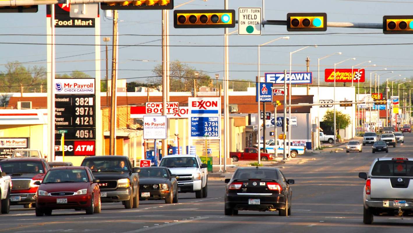 Traffic on a Texas street lined with various businesses and gas stations, featuring traffic lights overhead.