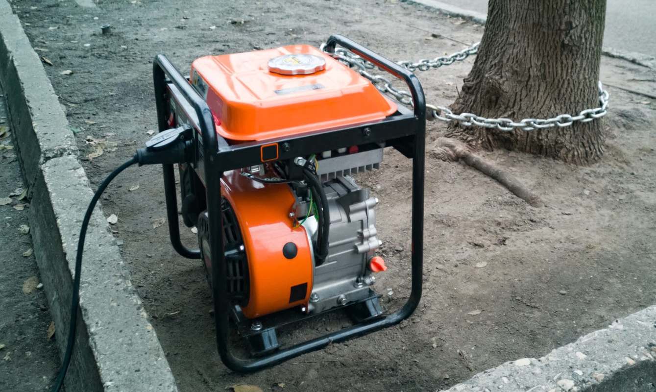 A portable generator is placed on the ground with a chain around it, near a tree and a concrete edge.