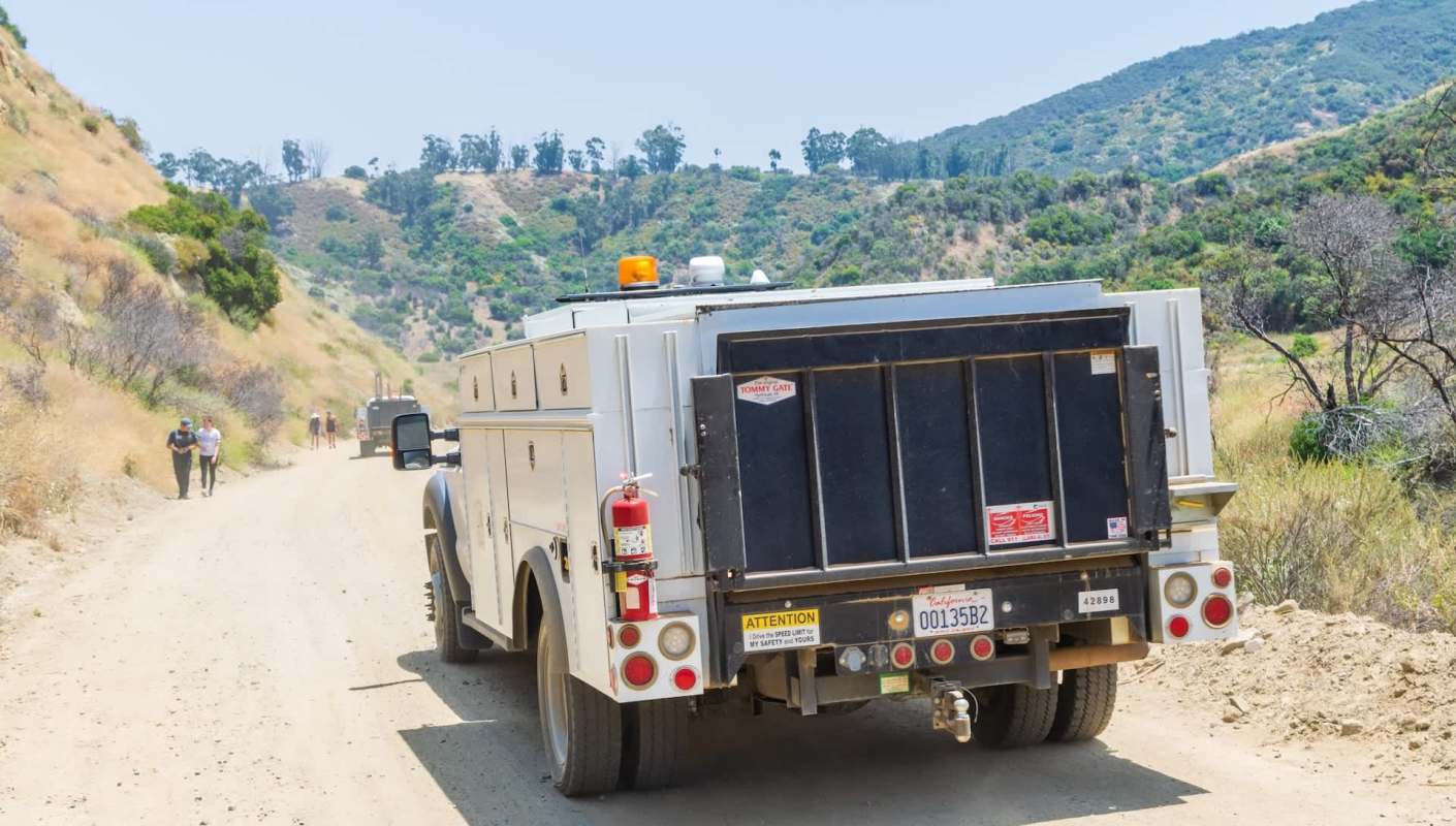 A white service truck is parked on a dirt road with hikers in the background and hills covered in vegetation.