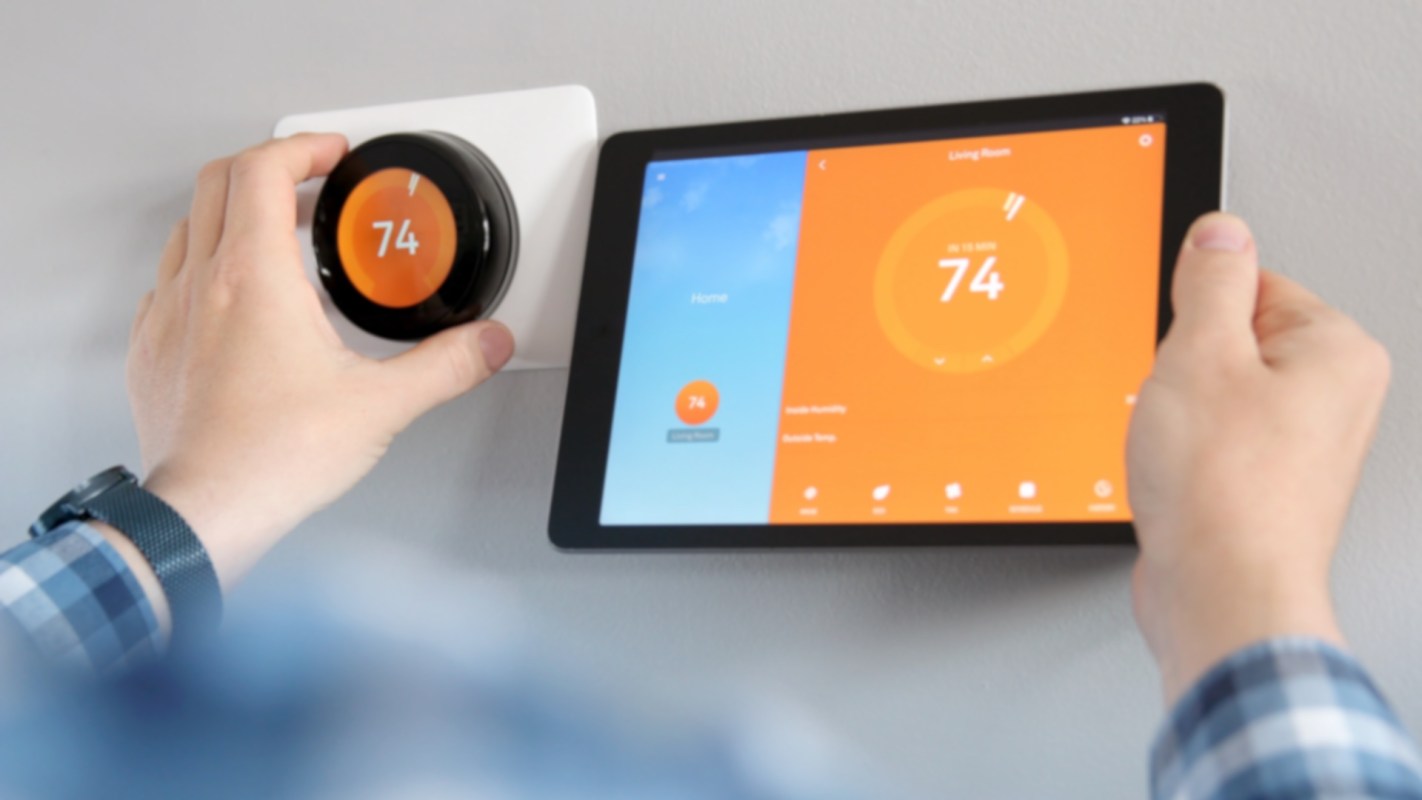 A person adjusts a smart thermostat while holding a tablet displaying temperature settings.
