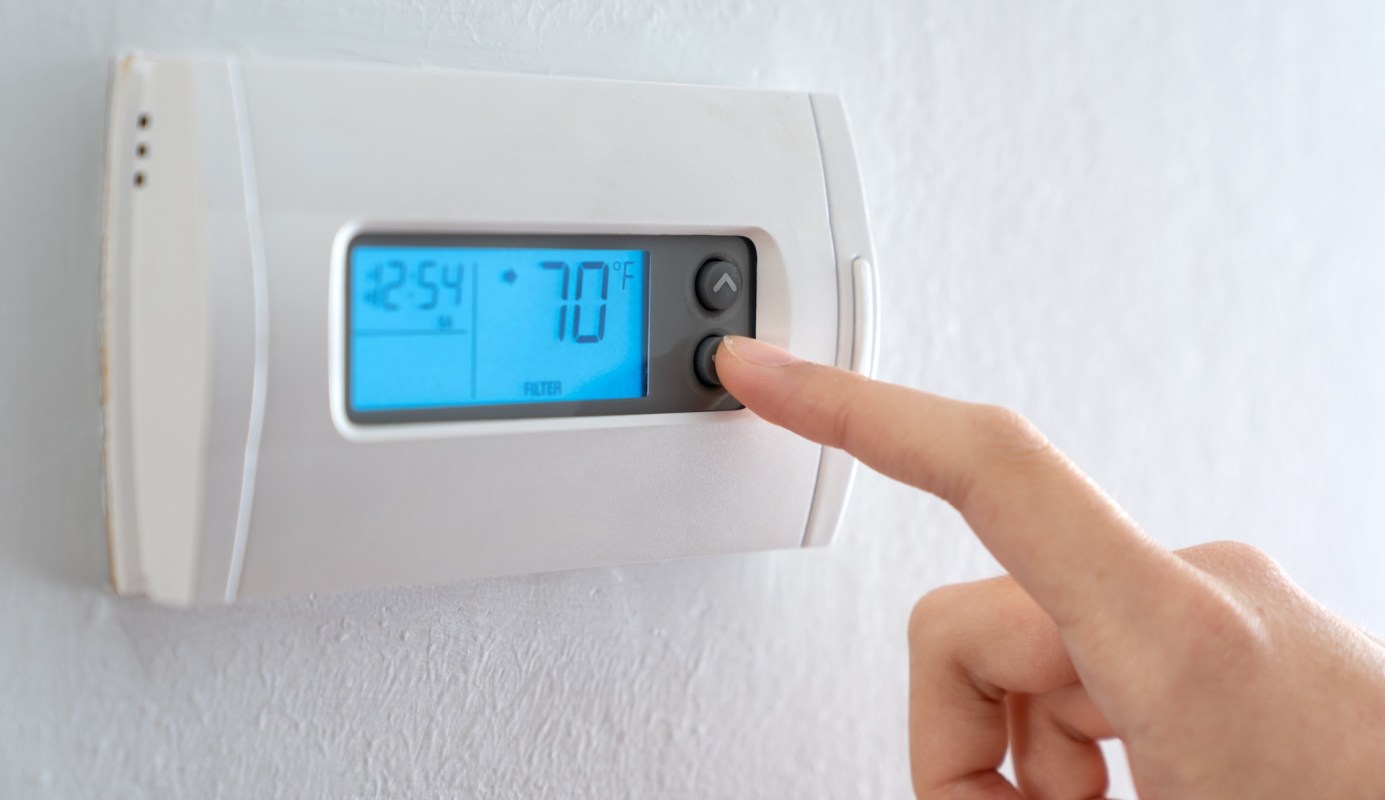 A hand adjusts the temperature on a digital thermostat displaying 70°F.