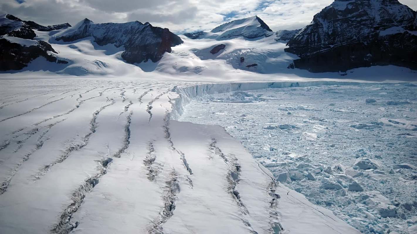A glacier with crevices and ice chunks, surrounded by snow-covered mountains under a cloudy sky.
