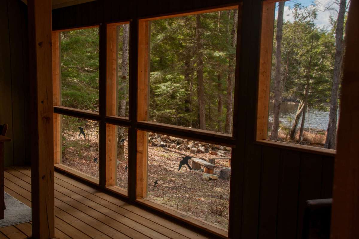 A view of a wooden cabin interior with large windows overlooking a forest and a river.