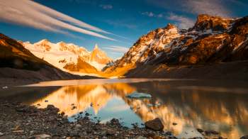 A mountain landscape reflecting in a lake at sunset, with snow-capped peaks and rocky shores.
