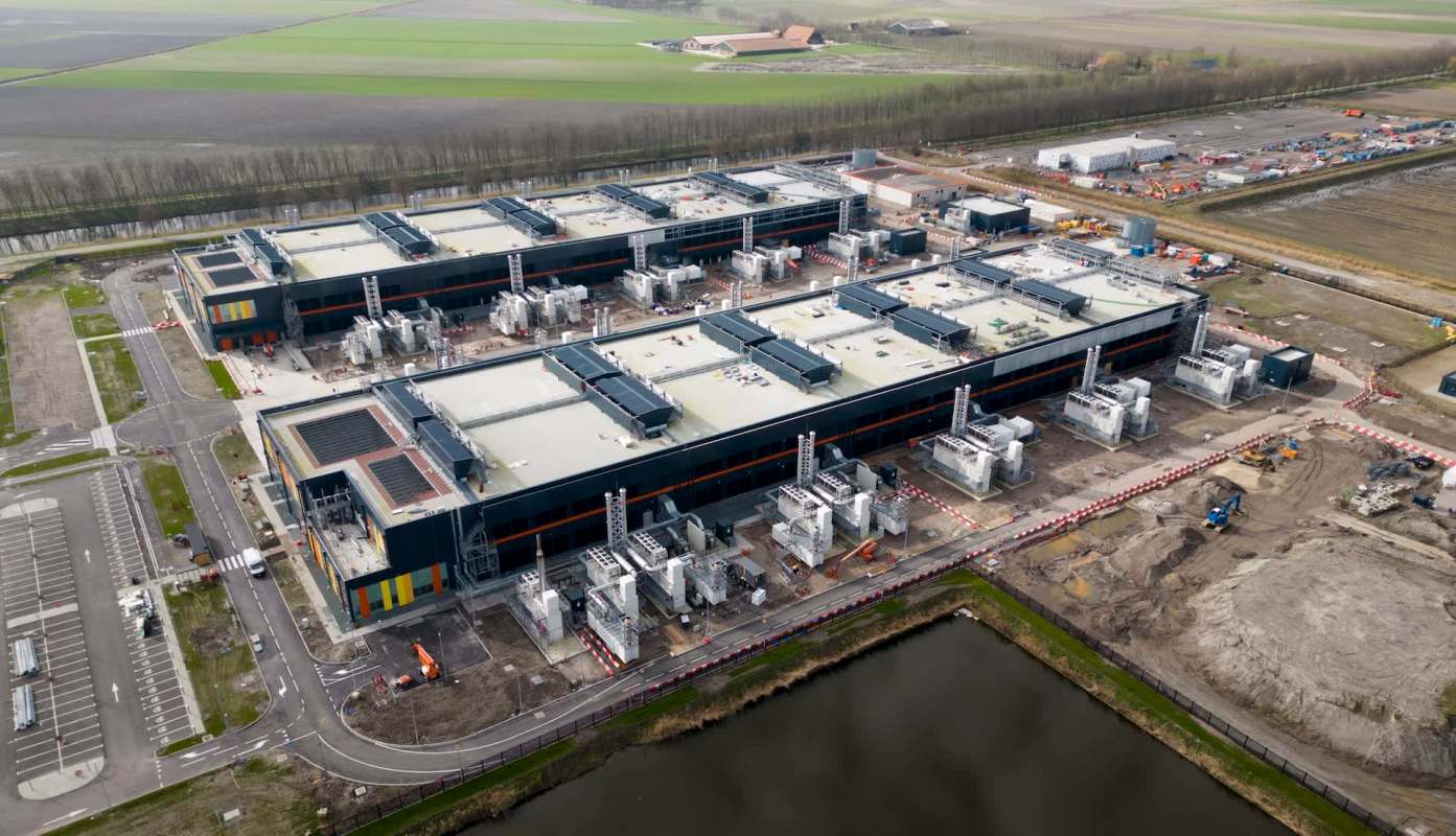 An industrial facility with multiple large structures and equipment, surrounded by fields and a waterway.