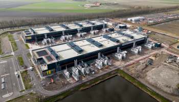 An industrial facility with multiple large structures and equipment, surrounded by fields and a waterway.