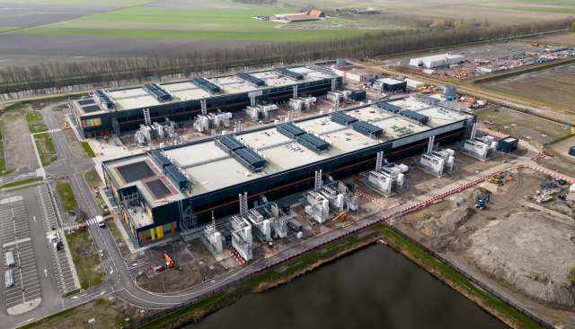 An industrial facility with multiple large structures and equipment, surrounded by fields and a waterway.