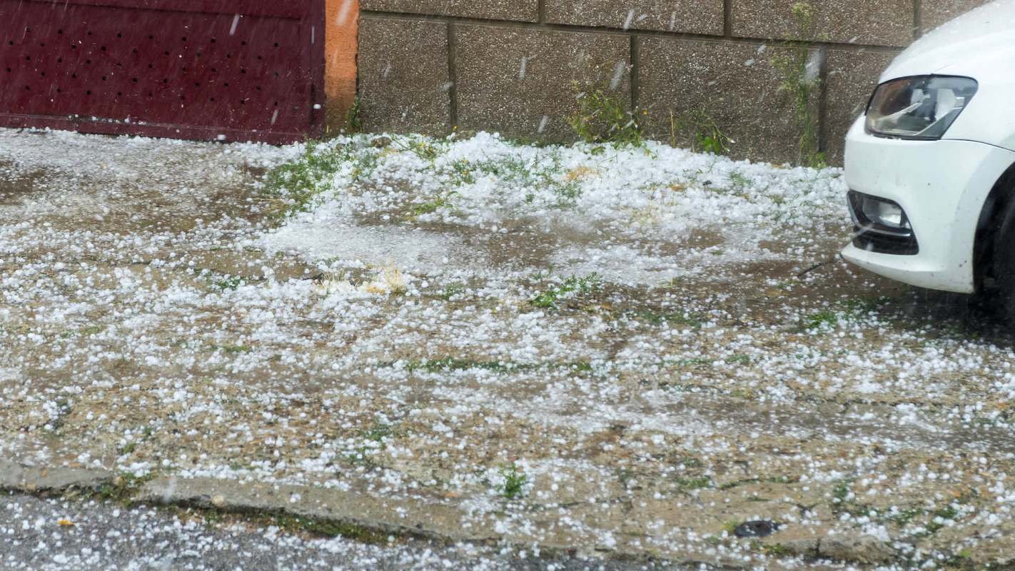 Hail covers the ground and a white car is parked on a flooded street.