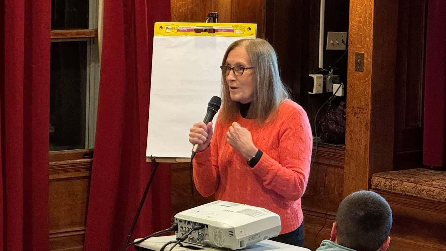 Massachusetts treasurer Deb Goldberg speaks into a microphone in front of a projector and a blank easel, with red curtains in the background.