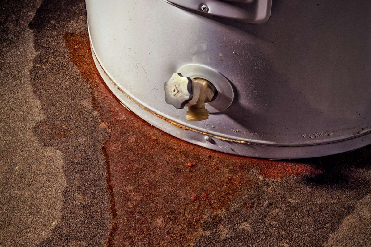 A white container with a valve leaks a reddish liquid onto a concrete surface.