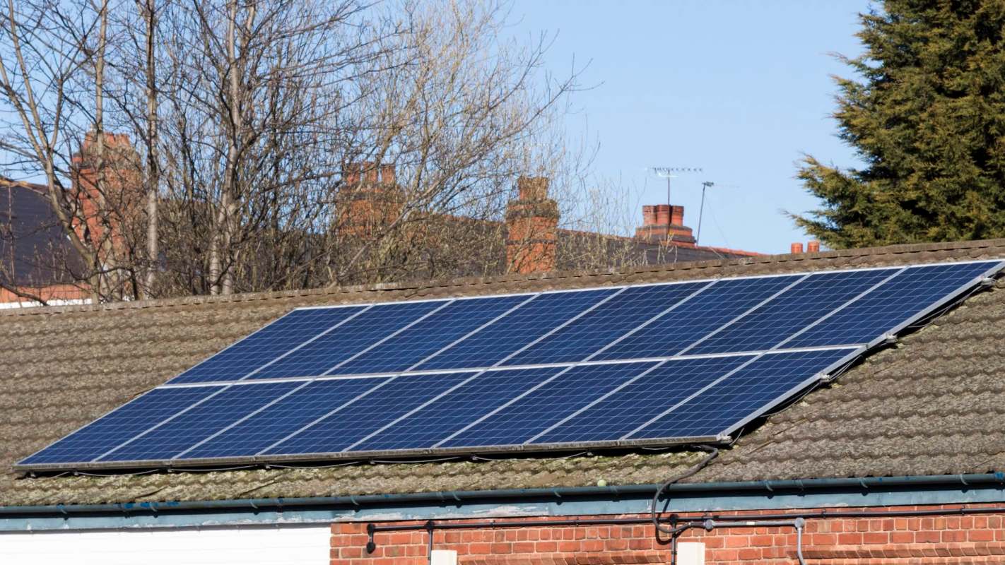 Solar panels installed on a sloped roof with surrounding trees and buildings in the background.