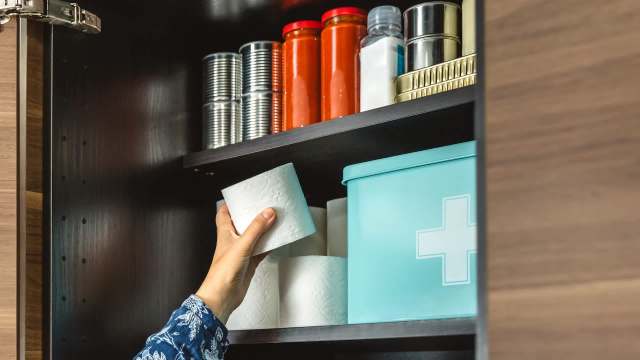 A hand holding a roll of toilet paper near a shelf with jars, cans, and a turquoise storage box.