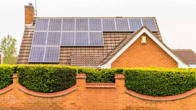 A house with solar panels on the roof and a brick wall topped with a hedge.