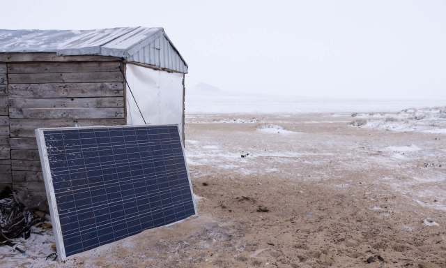 A solar panel is placed next to a small wooden structure in a snowy, cloudy landscape.