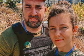 A man and a woman stand in a grassy area, both wearing tactical vests with identification badges.