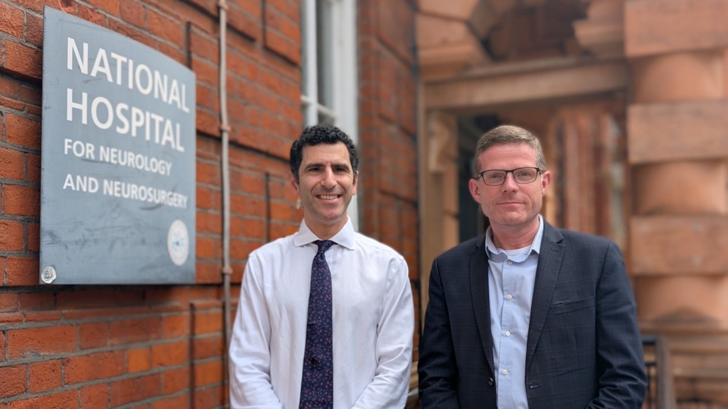 Two men stand outside the National Hospital for Neurology and Neurosurgery, beside a brick wall sign.