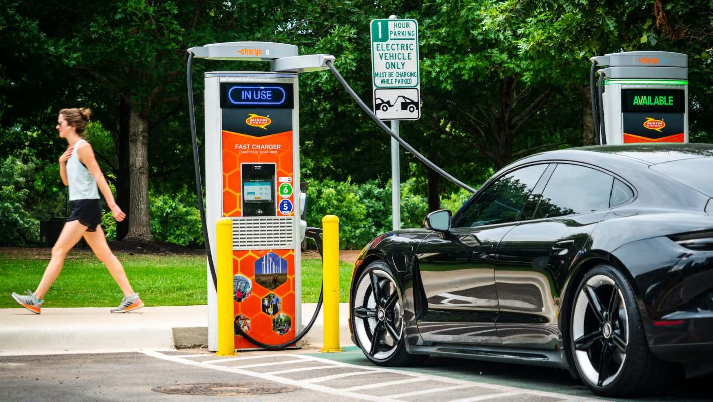 An electric vehicle charging station with a car parked nearby and a person walking in the background.