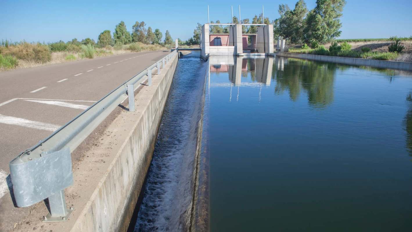 A concrete irrigation canal alongside a road, with a gate structure and trees in the background.