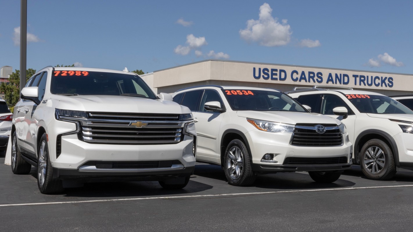 A row of used white SUVs parked in front of a building that says "USED CARS AND TRUCKS."