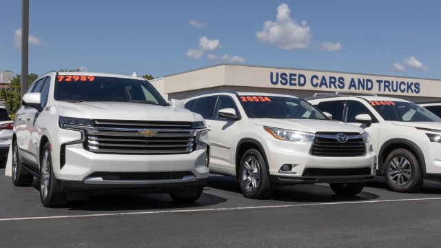 A row of used white SUVs parked in front of a building that says "USED CARS AND TRUCKS."