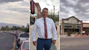 A man stands outside a gallery near a busy street with cars and a historic theater in the background.
