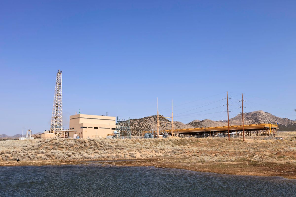 A site with a large building, a water body in the foreground, and mountains in the background under blue sky.