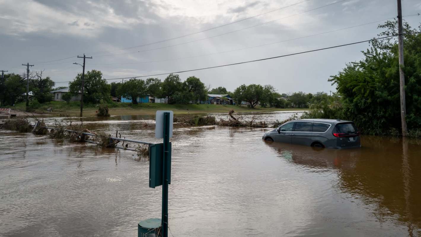 A flooded area with a submerged minivan and scattered debris in standing water, alongside utility poles and trees.