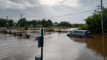 A flooded area with a submerged minivan and scattered debris in standing water, alongside utility poles and trees.