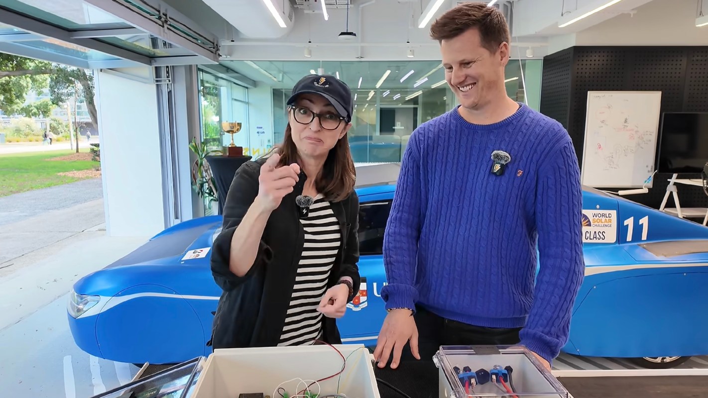 Two people stand in an indoor space with a blue solar car in the background, discussing a project with electronic components.