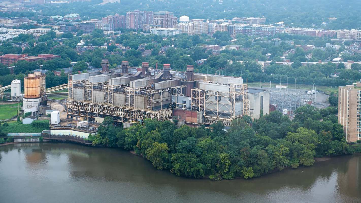 A large industrial building with smokestacks beside a river, surrounded by greenery and urban structures.