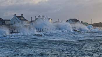 Waves crash against a barrier by coastal houses under a cloudy sky.