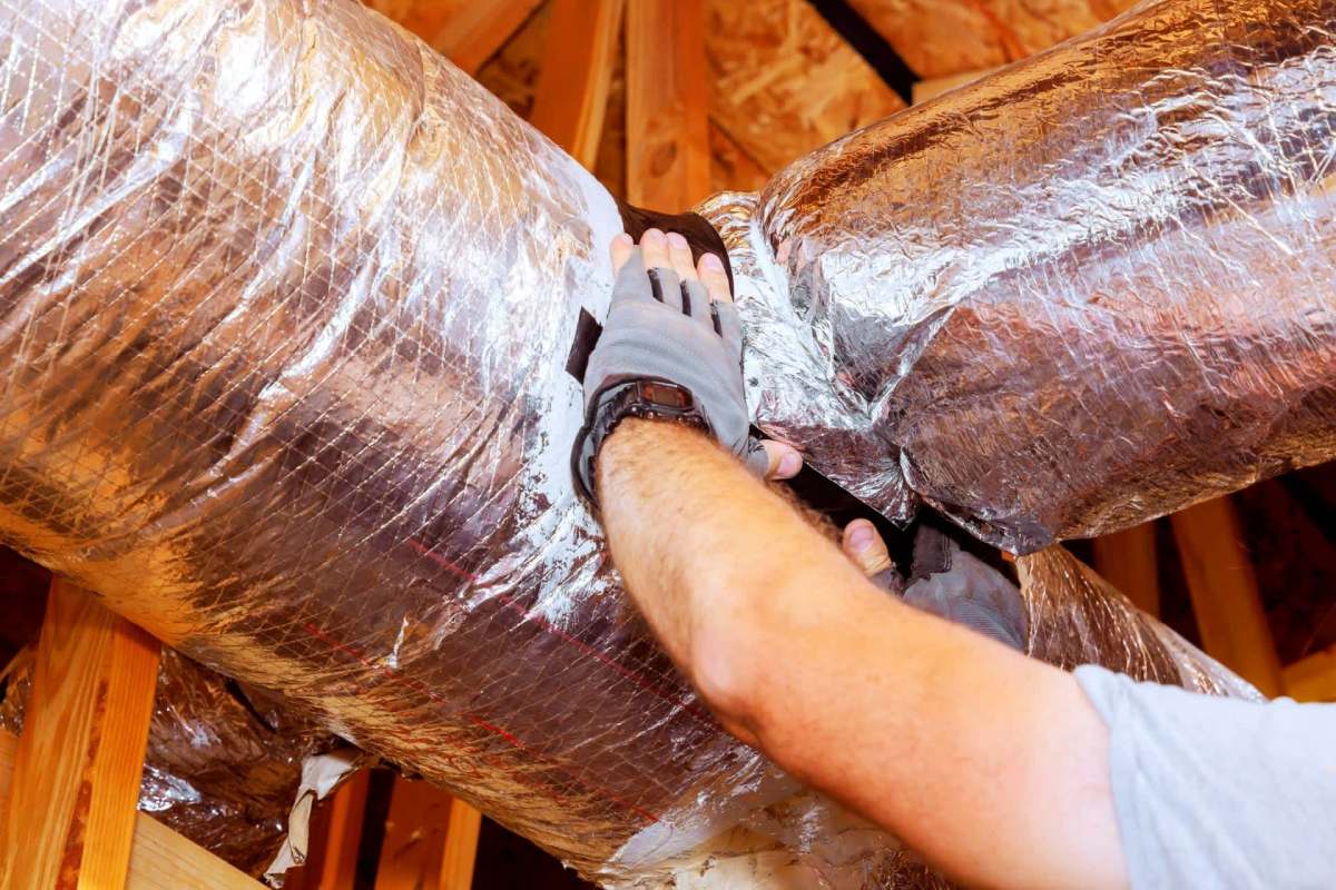 A person wearing gloves works on metallic ducting in an attic.