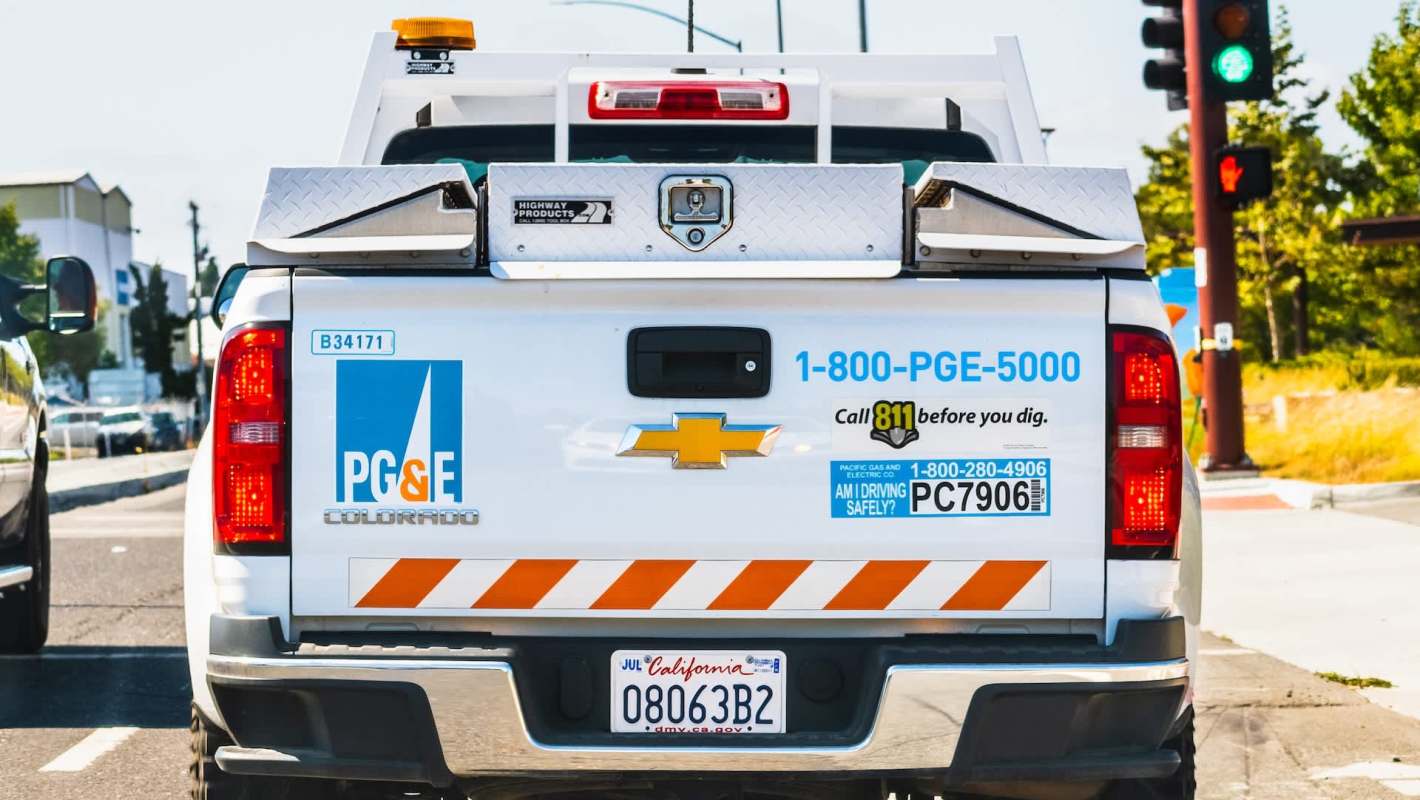 A utility truck with PG&E branding, parked at a traffic light, displaying a phone number and safety reminders.