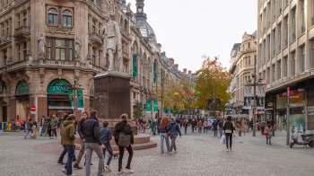 A busy street scene featuring pedestrians walking past shops and a statue in an urban area.