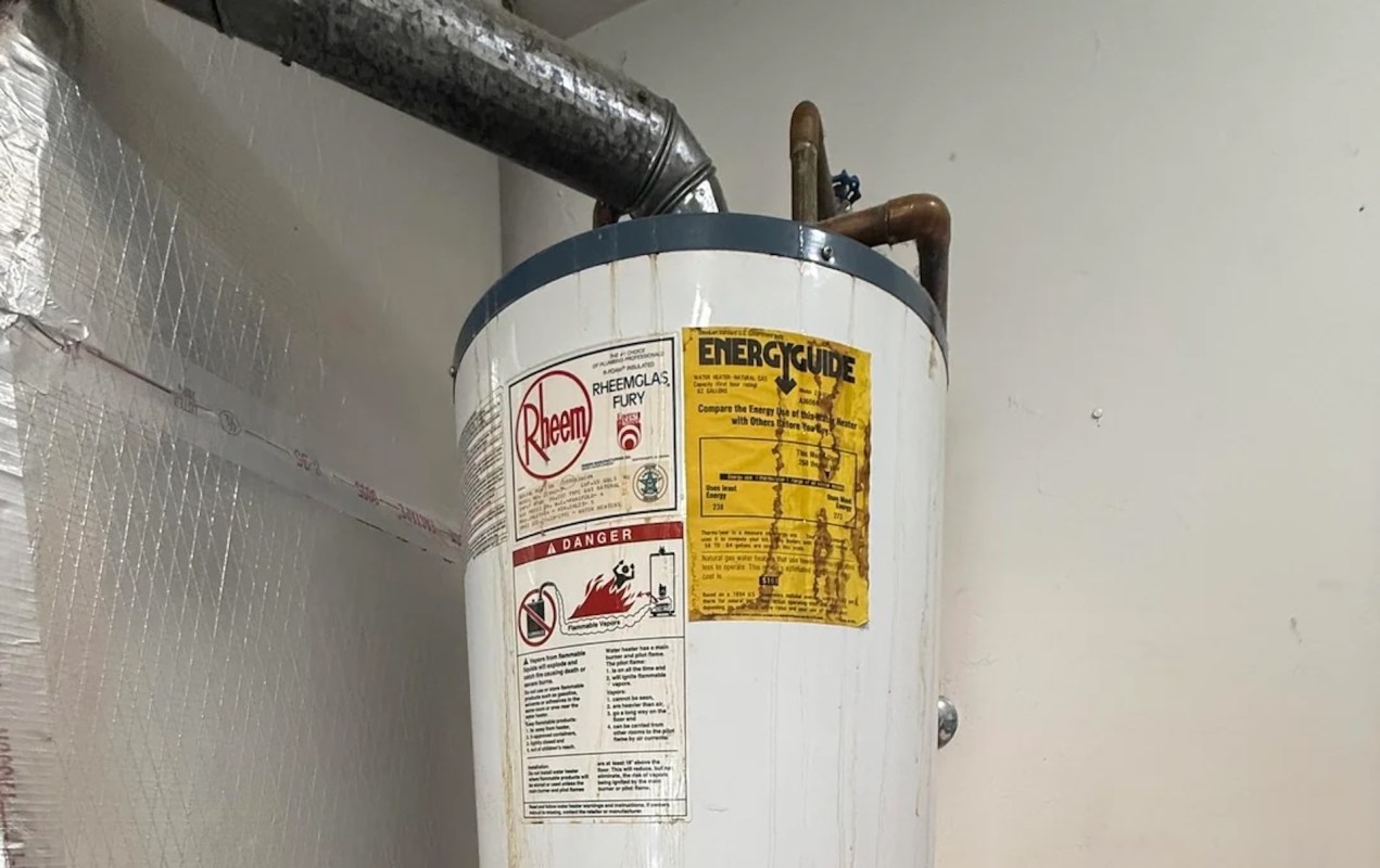 A water heater with warning labels and an energy guide sticker mounted on a wall, with rust stains running down the side.