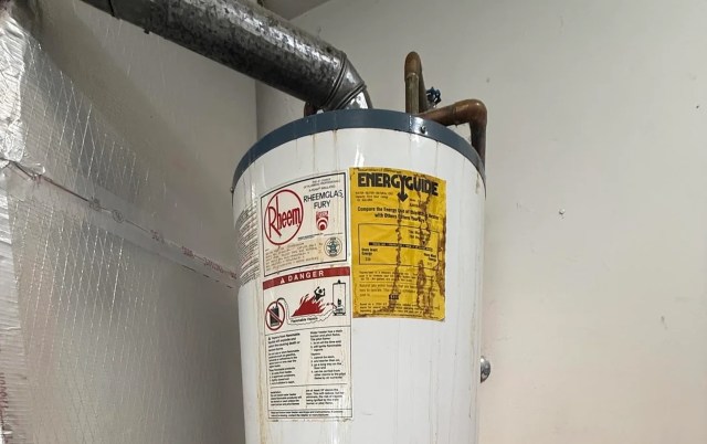 A water heater with warning labels and an energy guide sticker mounted on a wall, with rust stains running down the side.