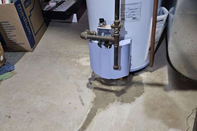 A water heater on a concrete floor with visible water stains and drainage pipes.